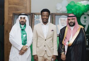 Three people in formal attire stand together at an event with Saudi Arabian decorations and green balloons in the background.