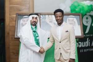 Two men wearing light colored formal attire shake hands at an event with green decorations and backdrop.