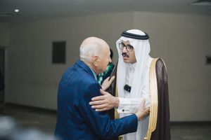 Two men greet each other warmly in a hallway, one wearing traditional Gulf Arab dress and the other in a blue suit.