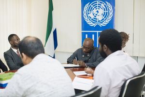 People attend a meeting with a United Nations flag visible in an office setting.