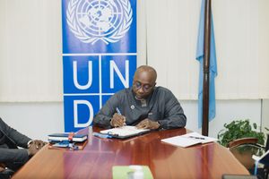 Someone sits at a desk reading documents with a UNDP logo and banner visible in the background during a meeting.