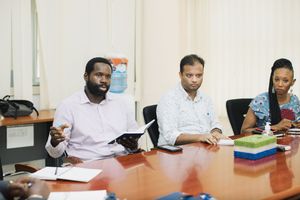 Three professionals participate in a business meeting around a wooden conference table in an office setting.