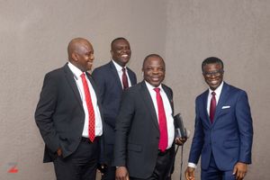 Four men wearing business suits and red ties standing together and smiling against a plain wall.