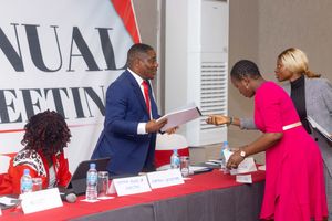 People in professional attire interact at a formal meeting with paperwork and signage visible in the background.
