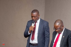 Two people in dark suits with red ties stand together, with one speaking into a microphone during a presentation.