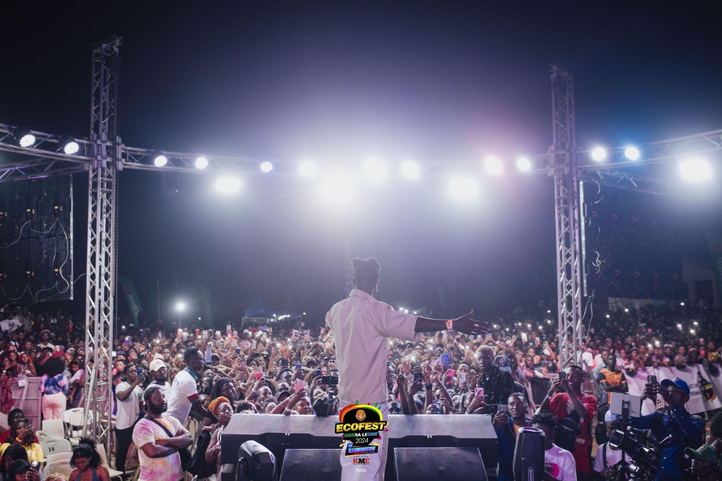 A performer on stage addresses a large nighttime crowd with bright stage lights illuminating the outdoor concert scene.