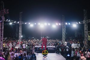 Performer in red outfit on stage at night performing to large crowd with bright stadium lighting and event scaffolding.