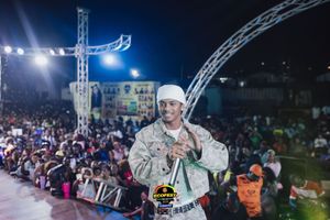 A performer in white attire on stage at a crowded outdoor nighttime concert venue with bright lights and metal rigging.