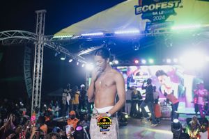 A shirtless performer energetically commands the stage at Ecofest with colorful lighting and stage equipment visible behind.