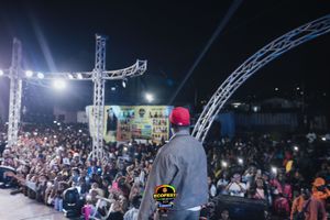 Concert performer on stage looks out at large nighttime crowd with lighting rigs and stage equipment visible.