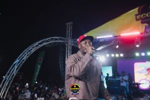 Performer in gray jacket holding microphone on stage under bright lights during nighttime concert performance.