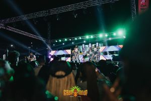 Concert stage setup with green lighting and video screens while audience watches performance from dark venue.