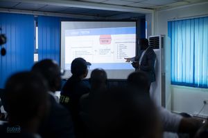 A presenter stands at the front of a darkened classroom with a presentation projected on a screen showing data.