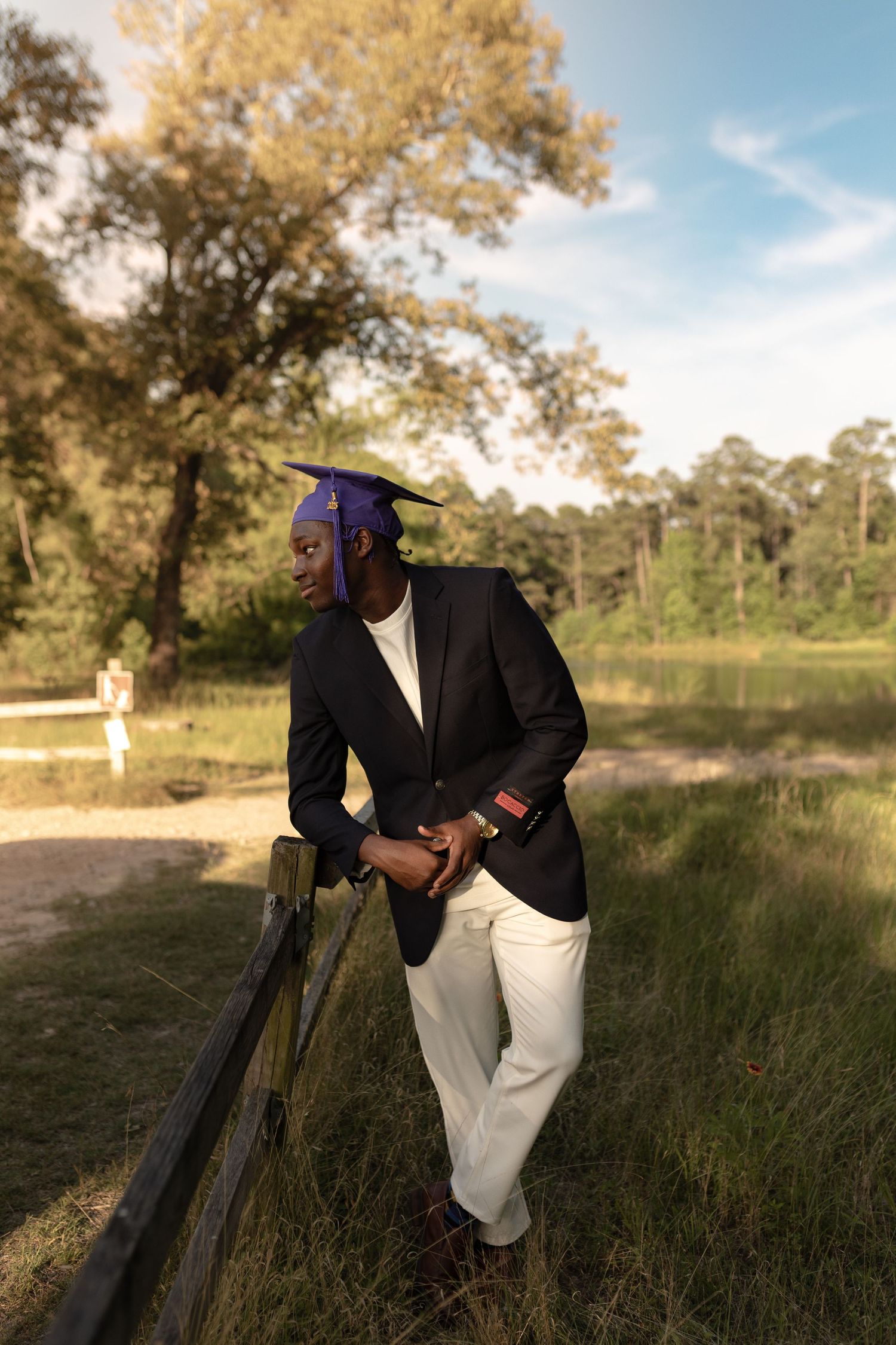 A person in all white clothing holds a basketball while standing on a dirt path in a wooded area on a sunny day.