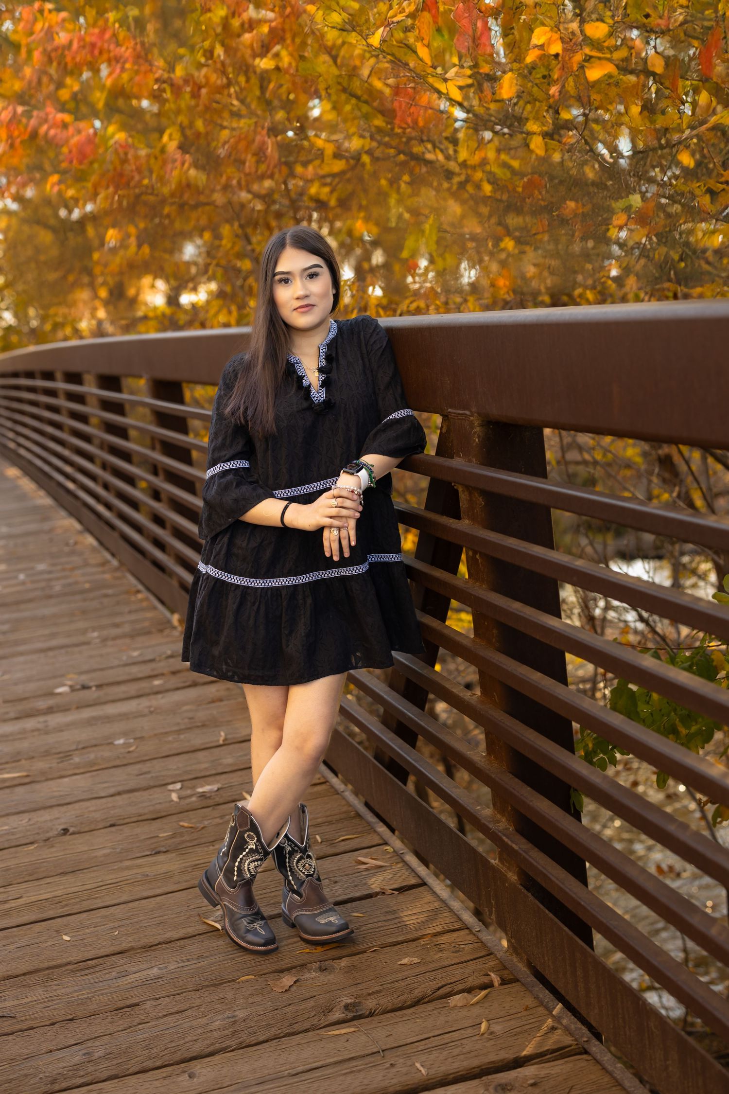Someone in a black dress and boots poses on a wooden bridge against vibrant autumn foliage in the background.