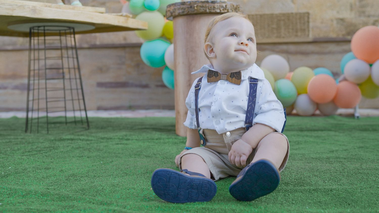 Baby in formal attire with bow tie and suspenders sitting on grass with balloons in background.