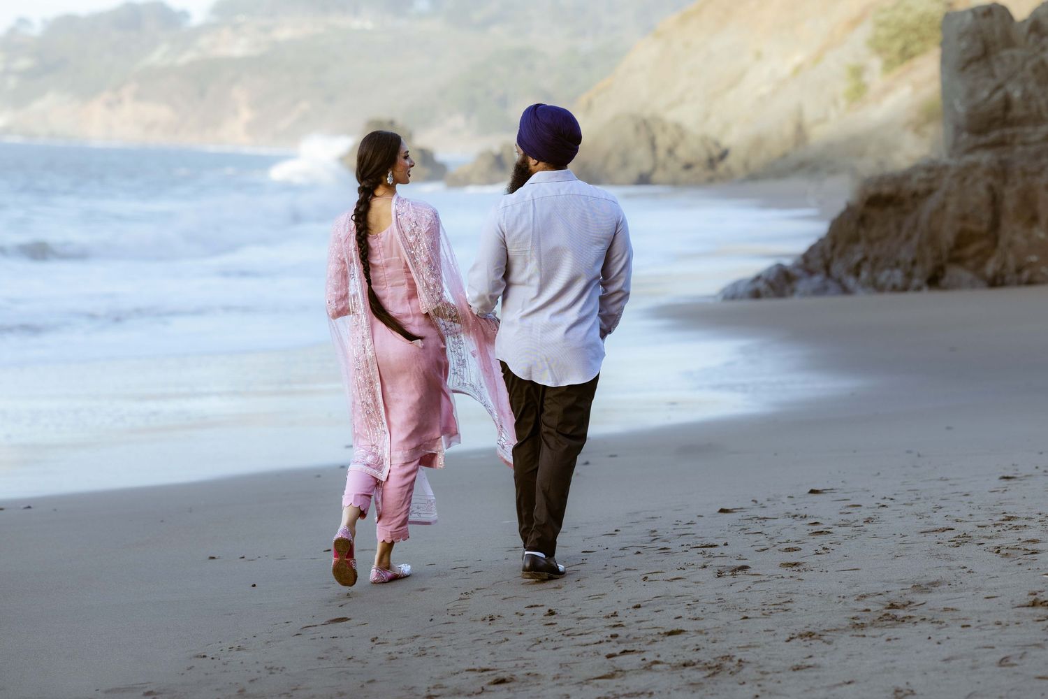 A couple wearing pink and blue outfits walks together along a scenic coastal beach with cliffs in the background.