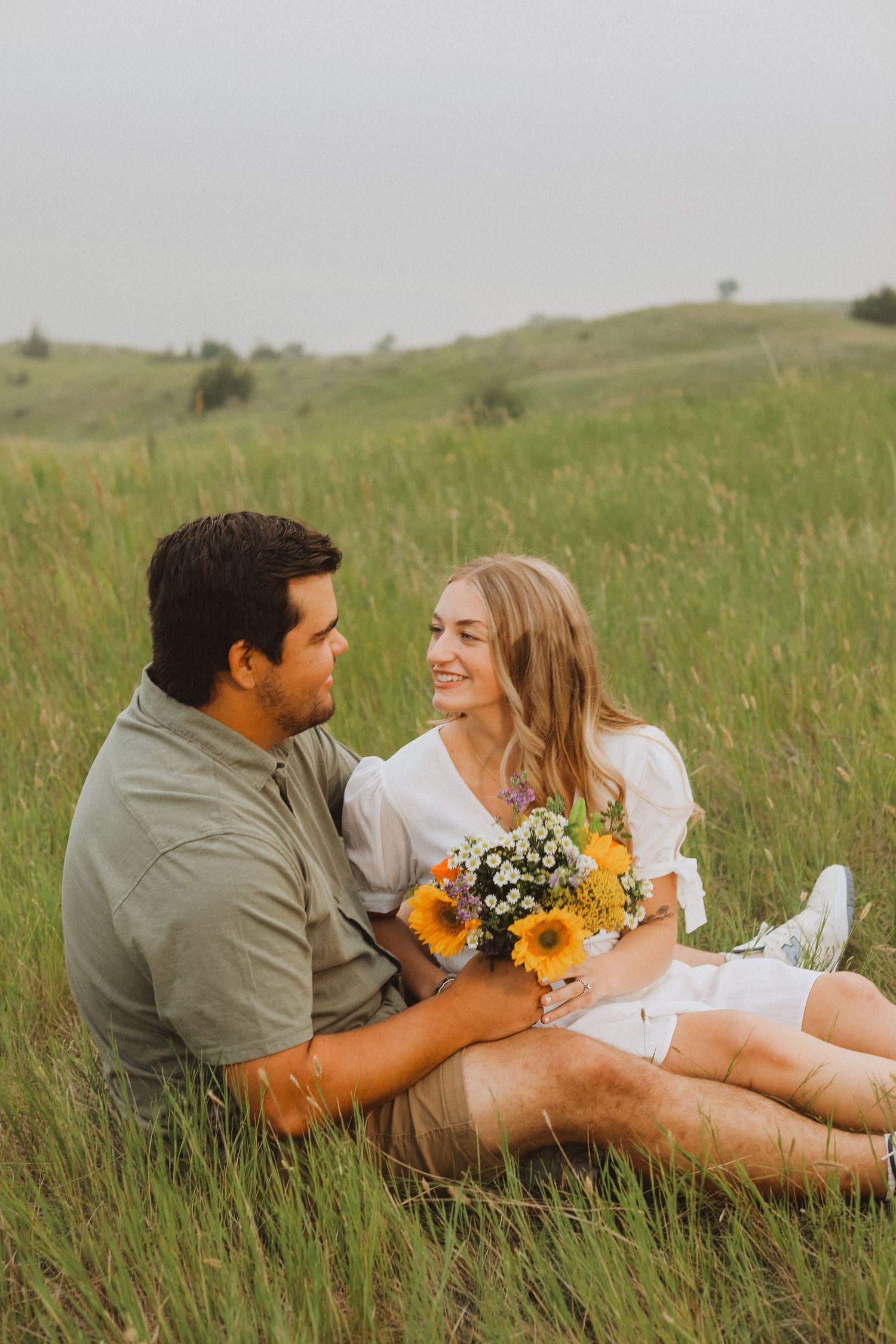 Couple sits in tall grass meadow sharing tender moment with sunflowers and wildflowers.