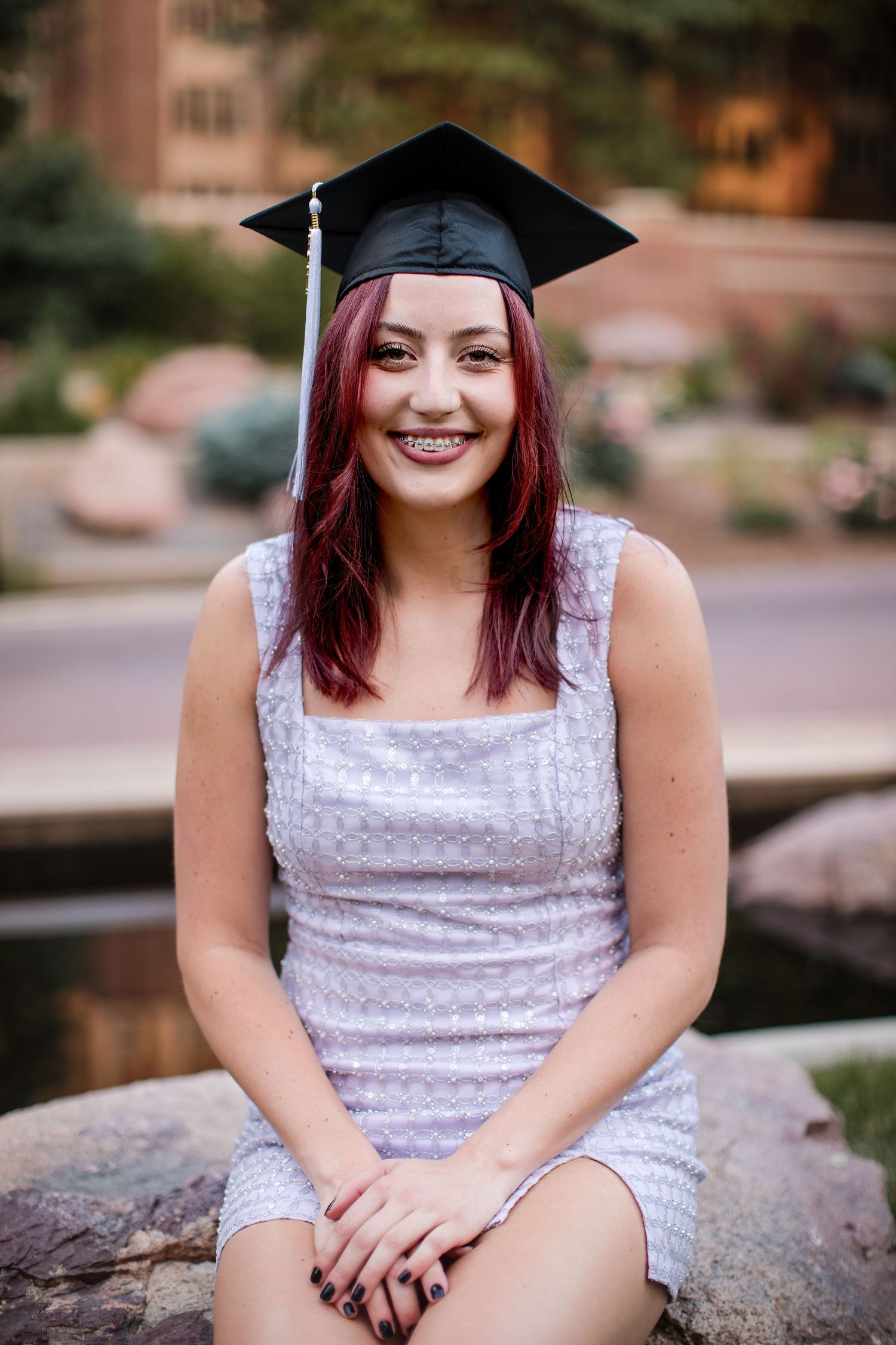 A graduate in a white dress and black mortarboard cap sits outdoors with red hair and a bright smile.