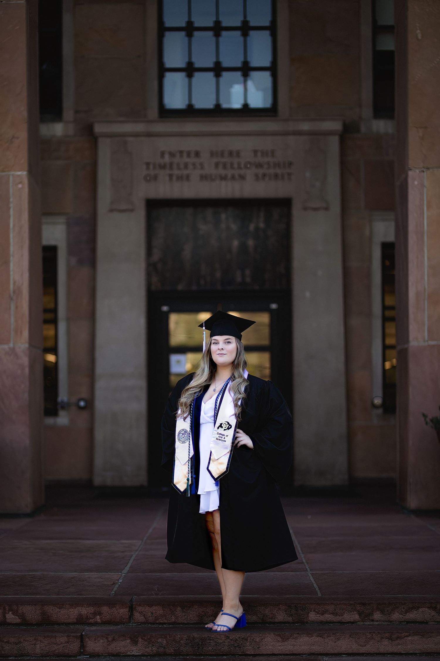 Graduate in traditional black cap and gown poses on stone steps in front of university building entrance.