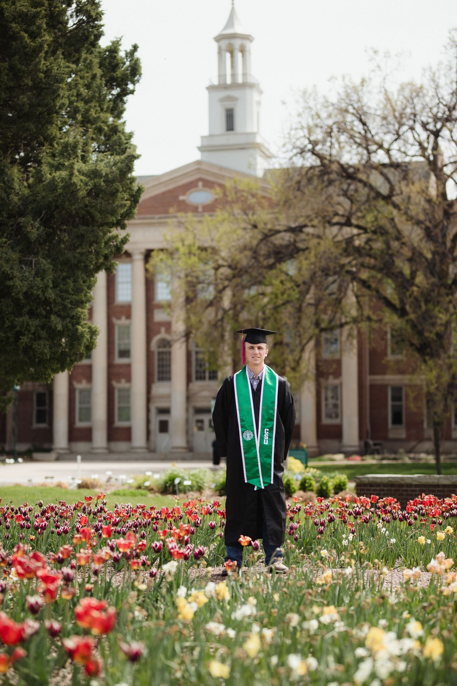 Graduate in traditional cap and gown with green sash stands in field of colorful tulips in front of stately brick building.