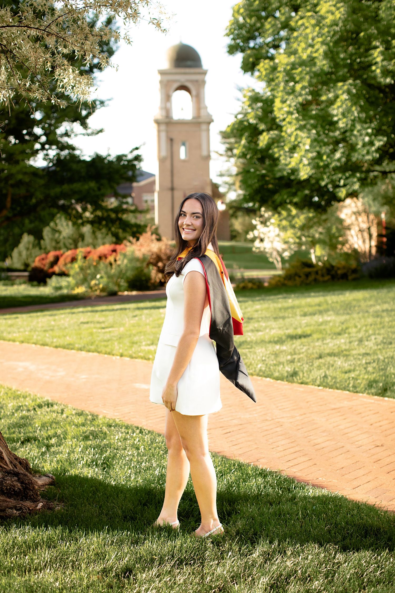 Student in white dress stands on brick path near campus bell tower on sunny summer afternoon.