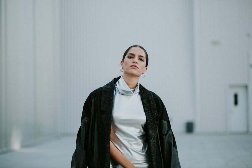 Young woman in black leather jacket and silver turtleneck posing confidently outdoors.