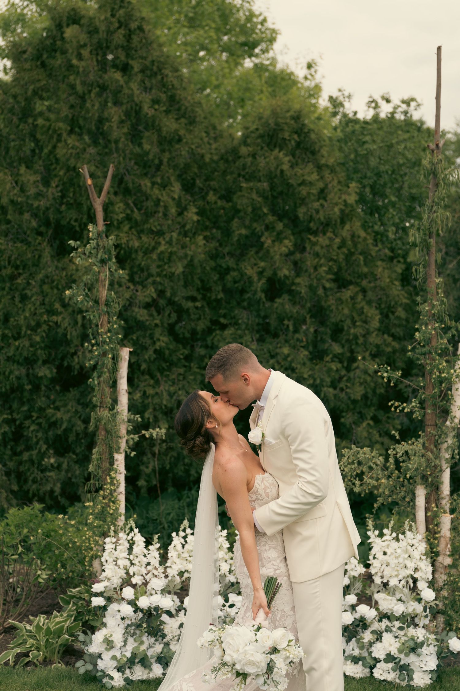 A romantic moment shared between a couple in white attire surrounded by delphinium flowers in a garden setting.