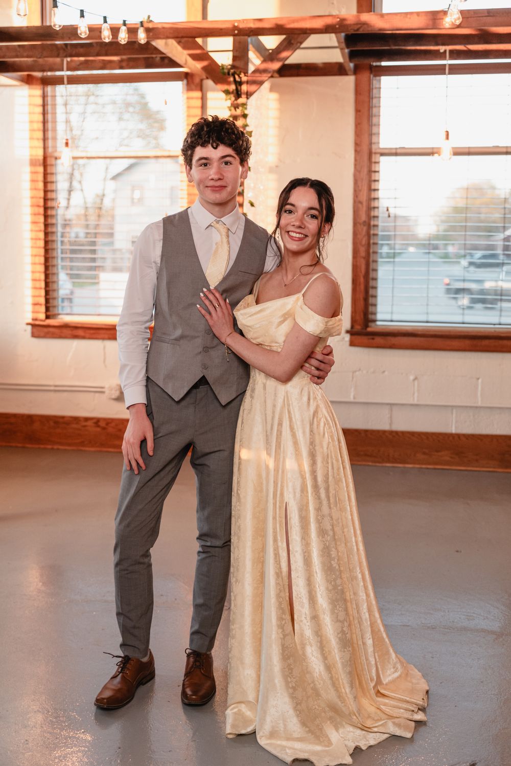 A couple poses at a formal event, with the person in grey standing next to someone in a champagne-colored flowing dress.