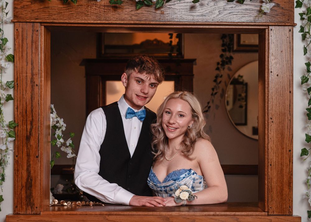 A couple poses together in formal attire by a wooden doorframe decorated with greenery at a special event.