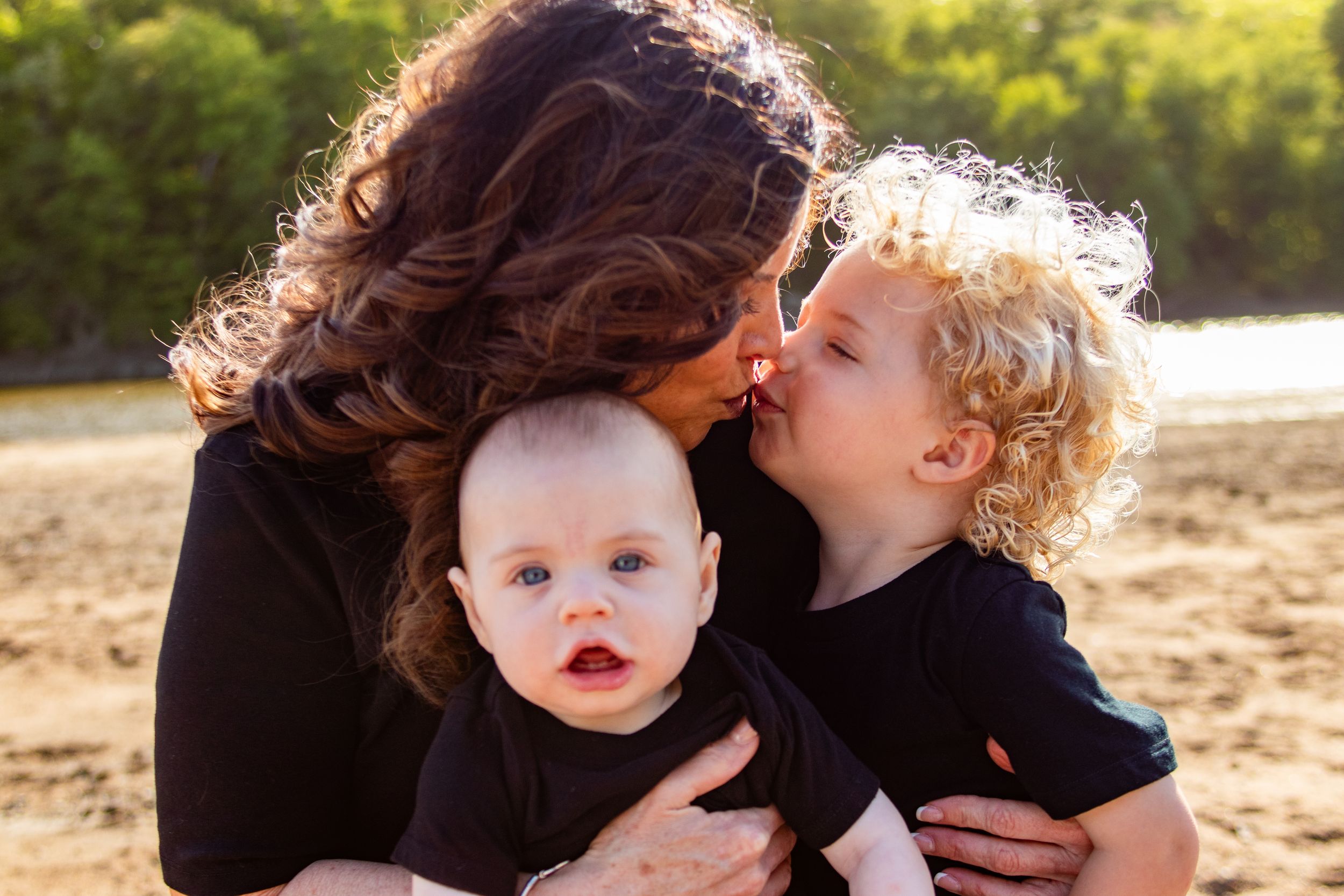 A mother holds her baby while her curly-haired toddler kisses her cheek outdoors on a sunny day.