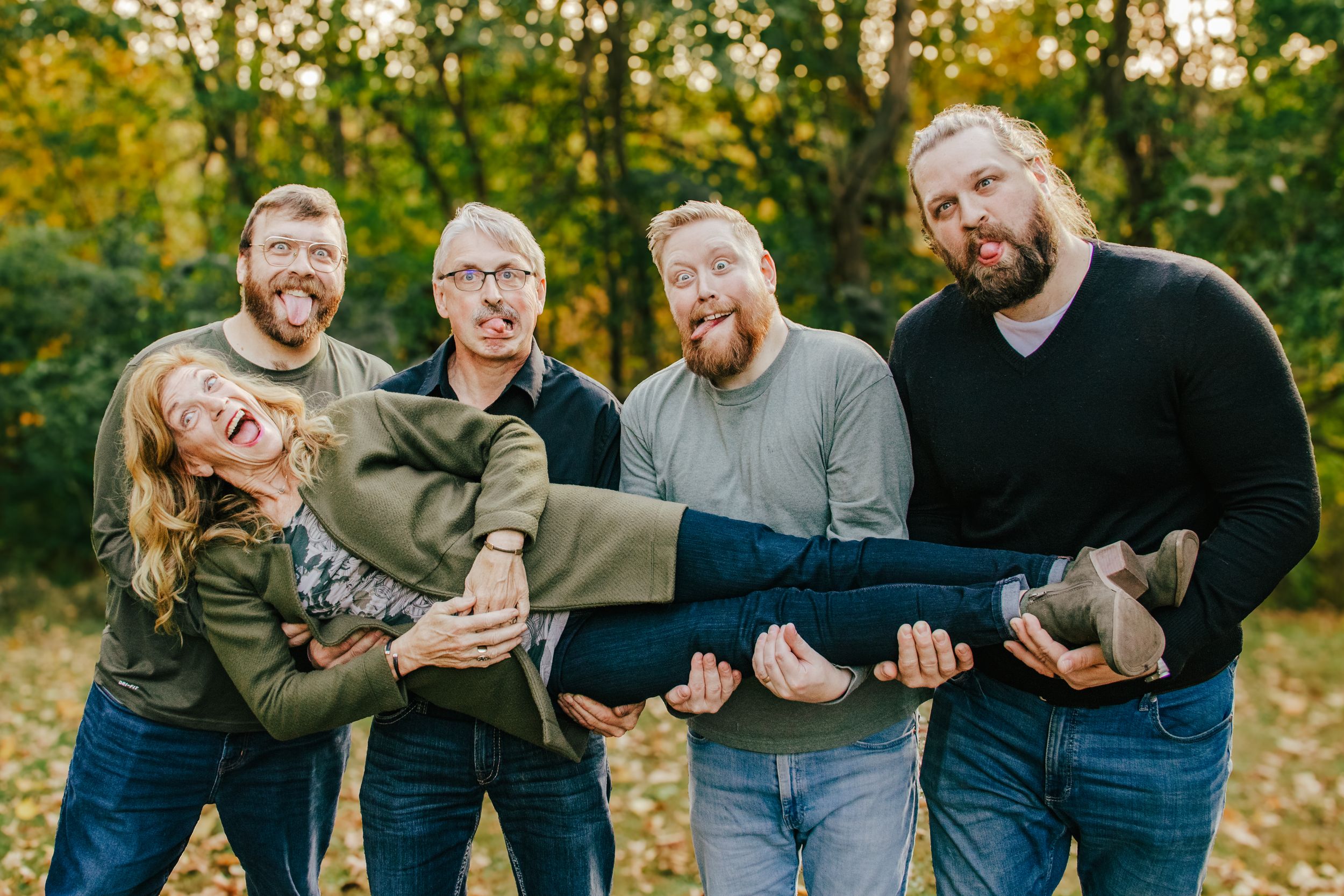 Five friends pose playfully outdoors in fall foliage, with four men lifting a laughing woman horizontally.