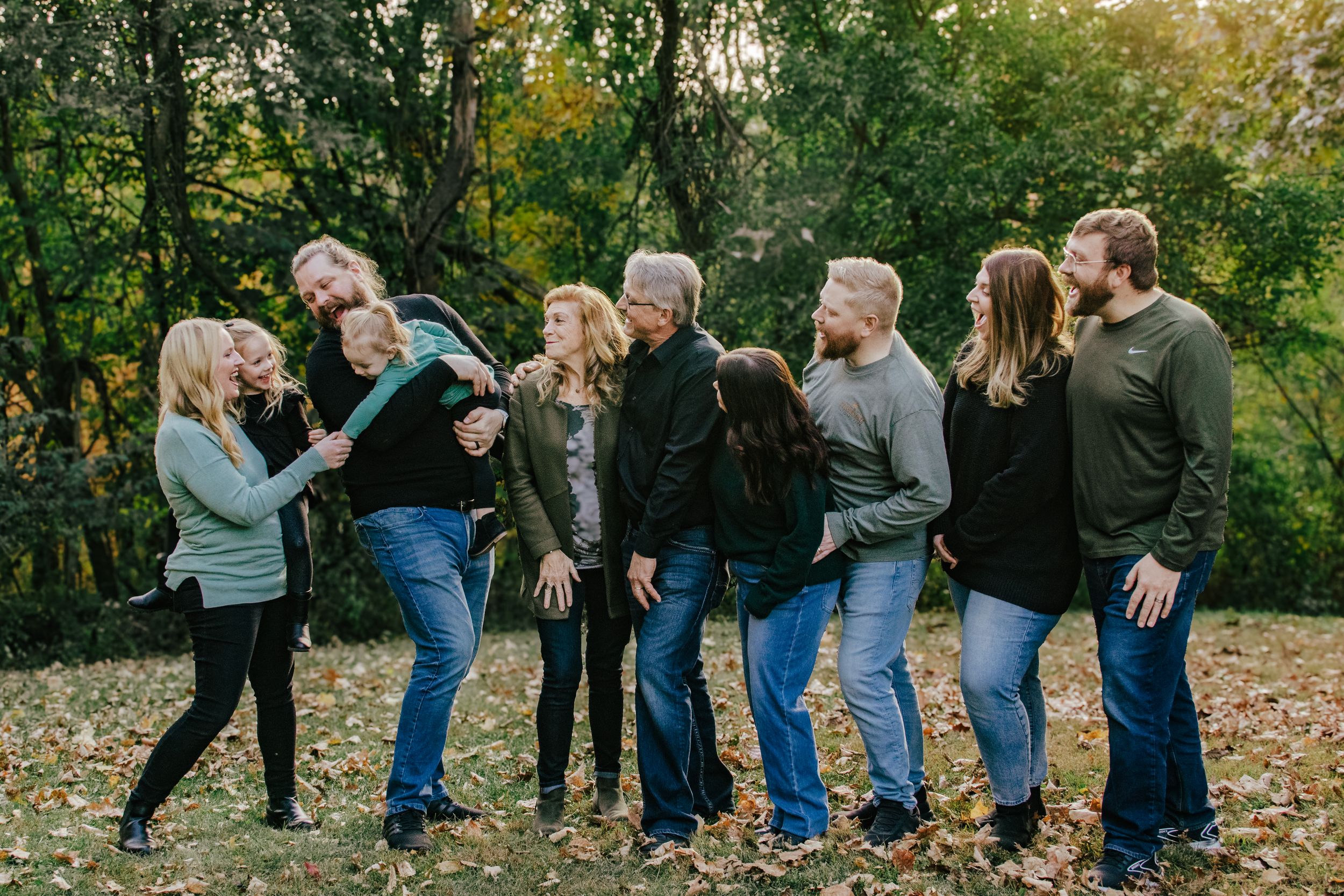 Large family group laughing together outdoors in autumn park with colorful fall foliage backdrop.