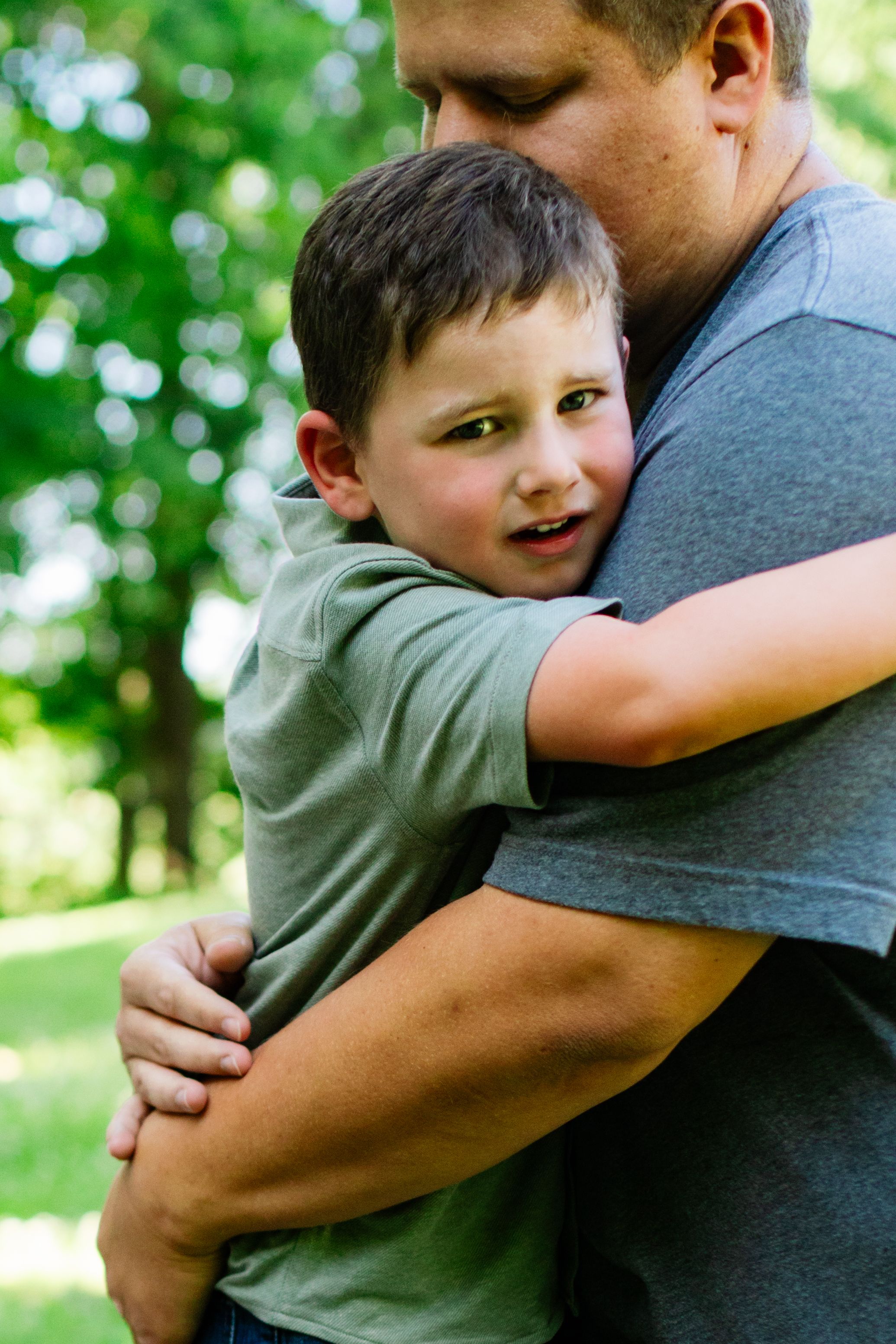 A young boy hugs an adult man outdoors in a green park, both wearing gray shirts in a warm embrace.