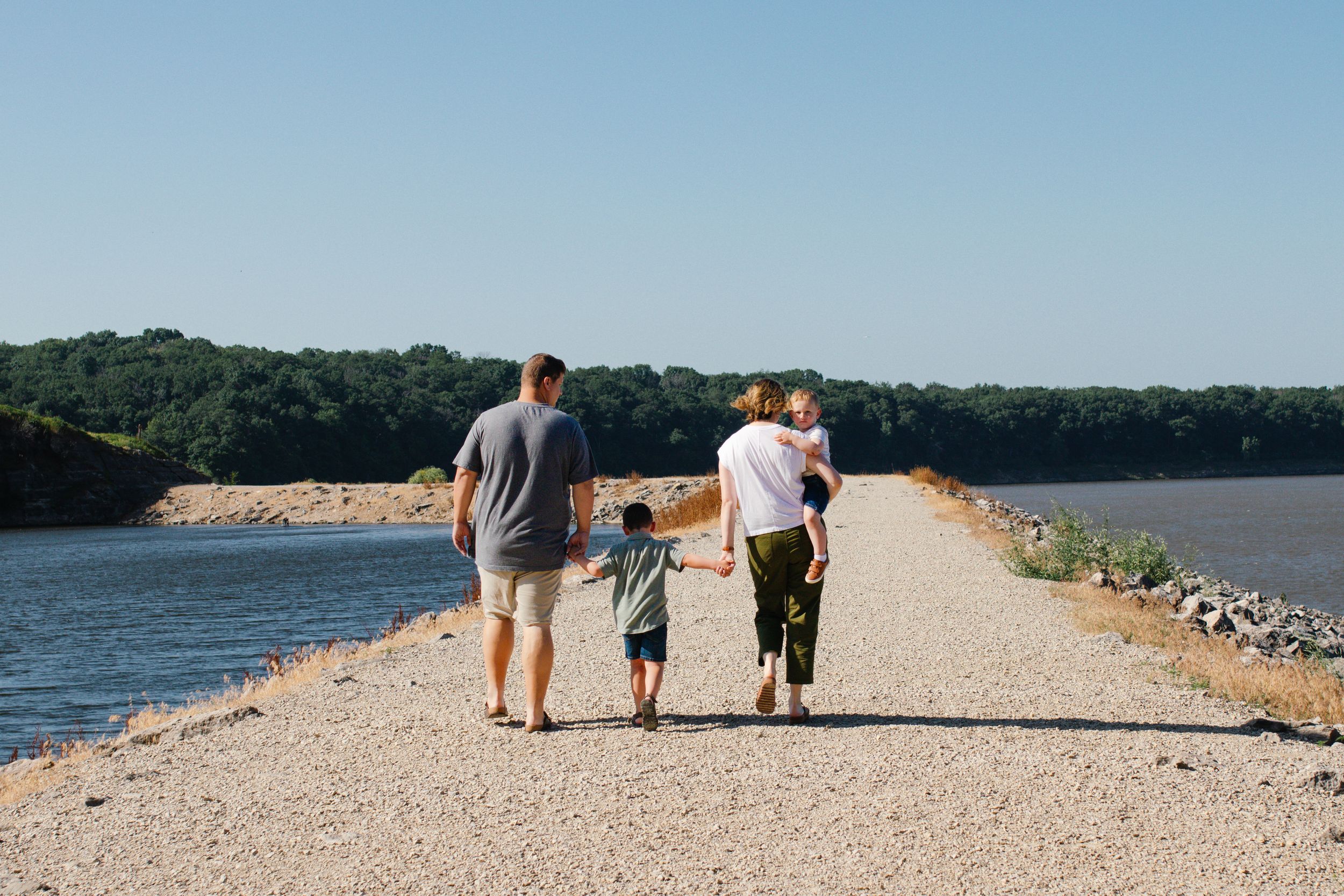 Family of four walking along a scenic gravel path beside a calm lake surrounded by lush green trees.