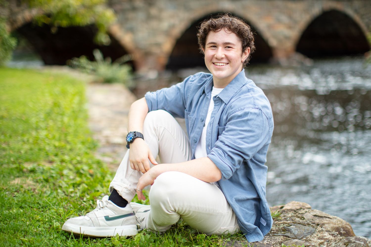 Student relaxes outdoors on stone ledge near flowing stream wearing casual denim shirt and khaki pants.