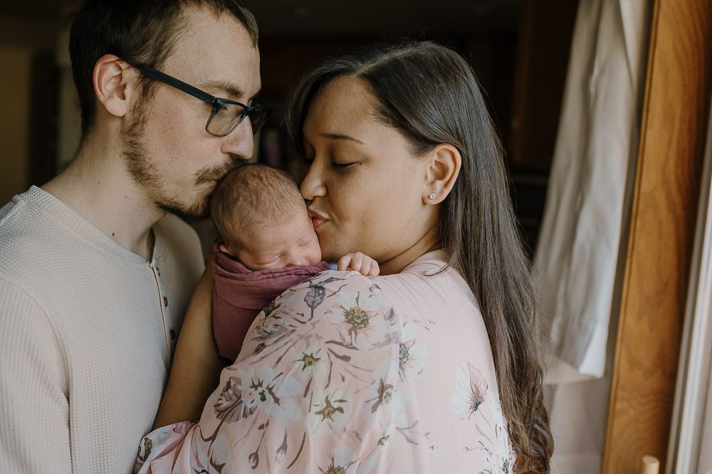 mom and dad cuddle close and kiss newborn baby who lies on mom's shoulder during newborn pictures