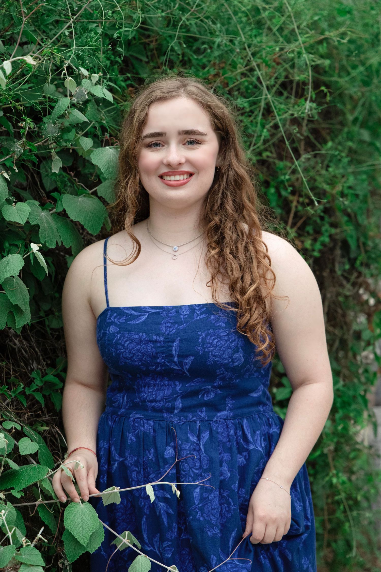 Person in a royal blue sleeveless dress poses outdoors against lush green foliage on a sunny day.