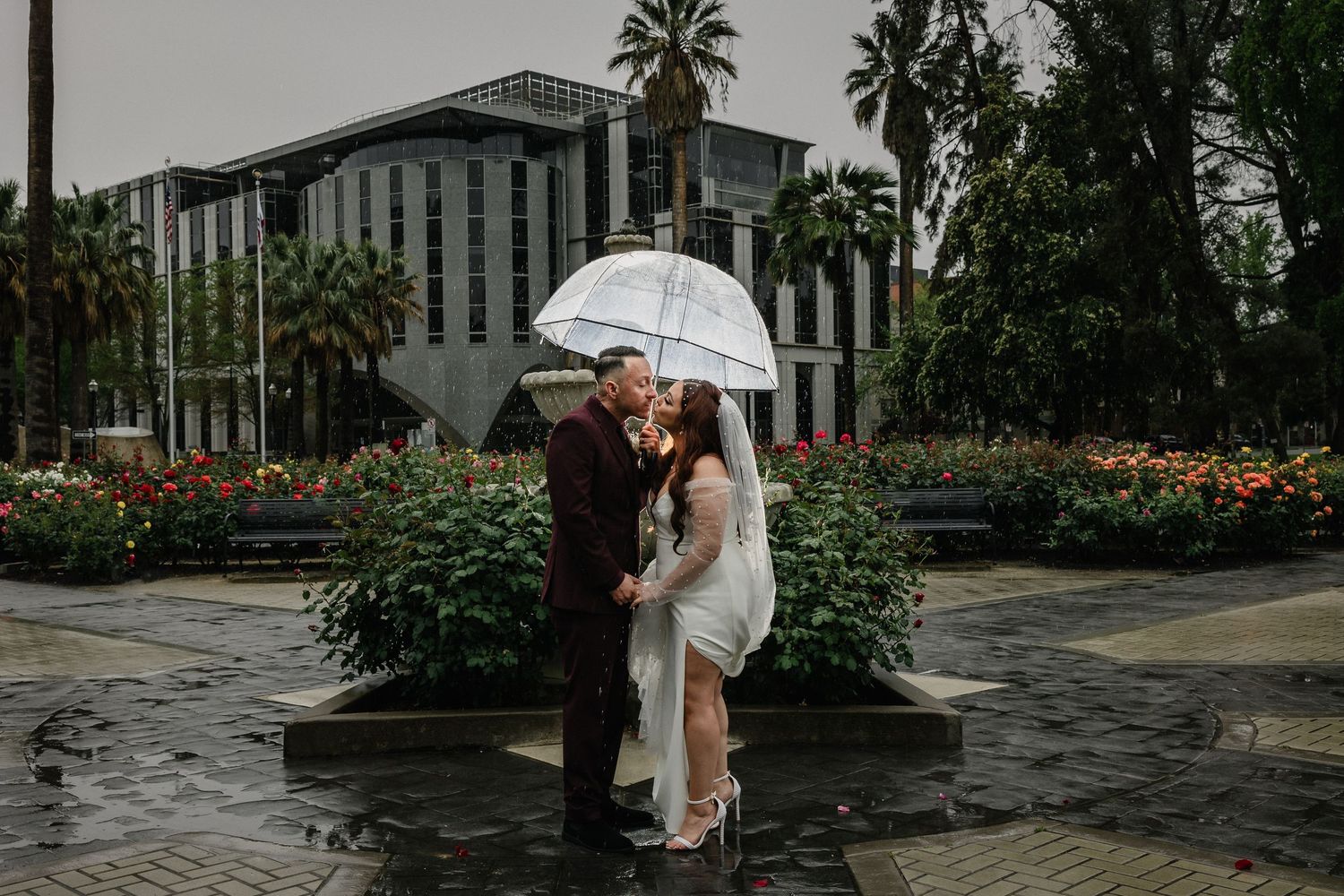 A wedding couple shares a romantic moment under a white umbrella in a rainy garden with flowers and a building in the background.