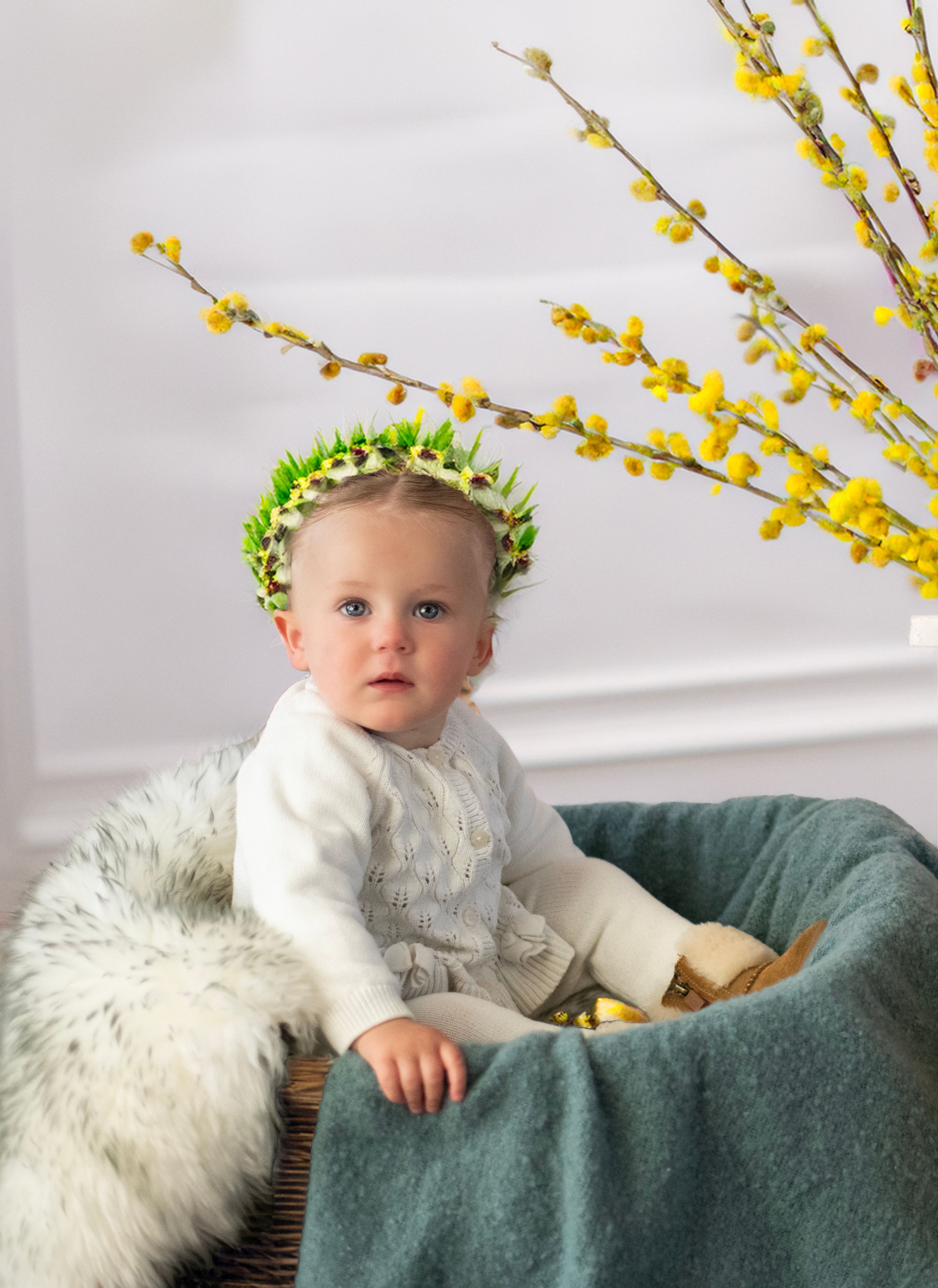 Young child in white sweater wearing floral crown sits in teal chair with spring forsythia branches in background.