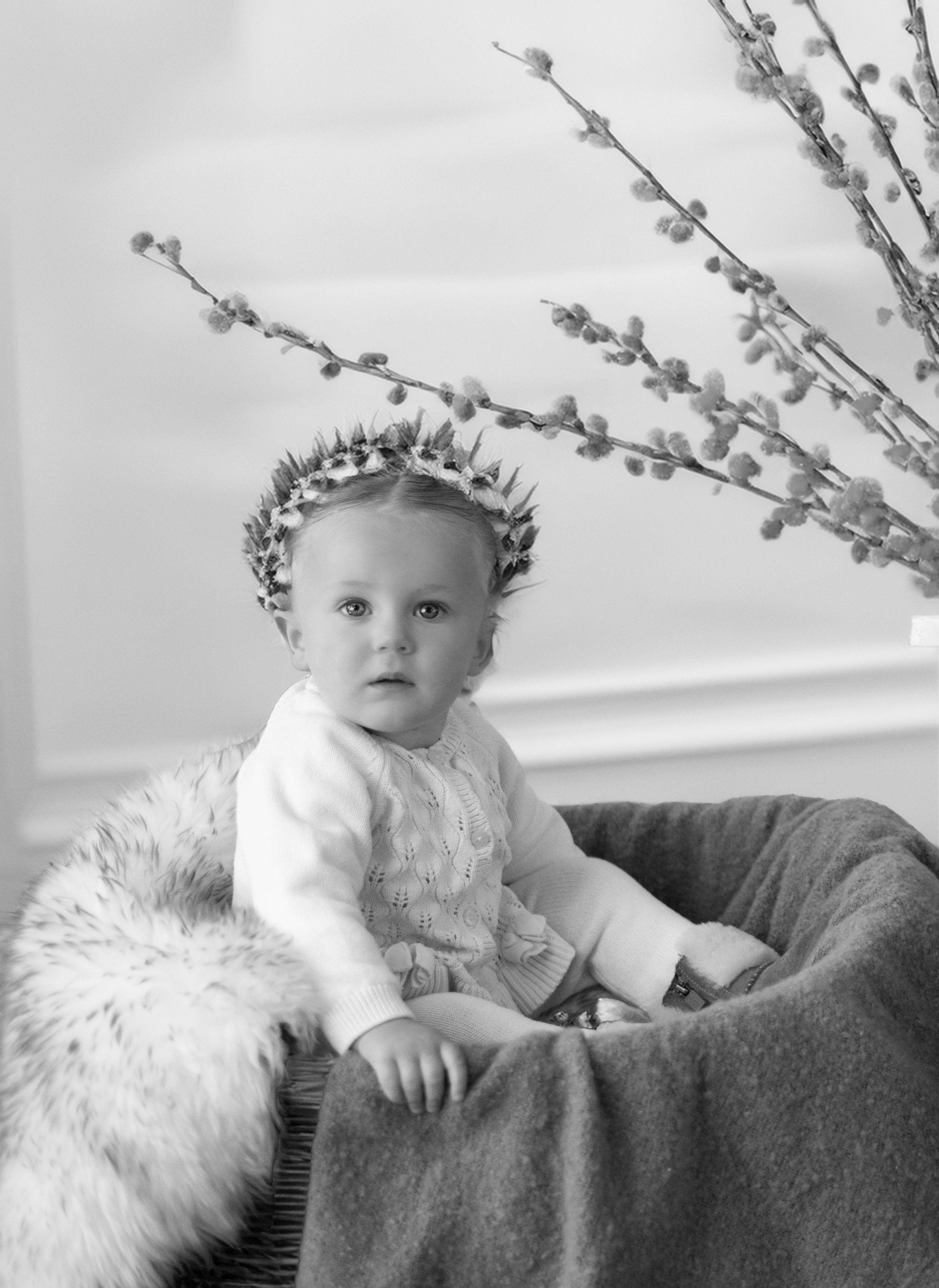 Black and white photograph of a small child wearing a crown sitting in a plush chair with willow branches behind them.