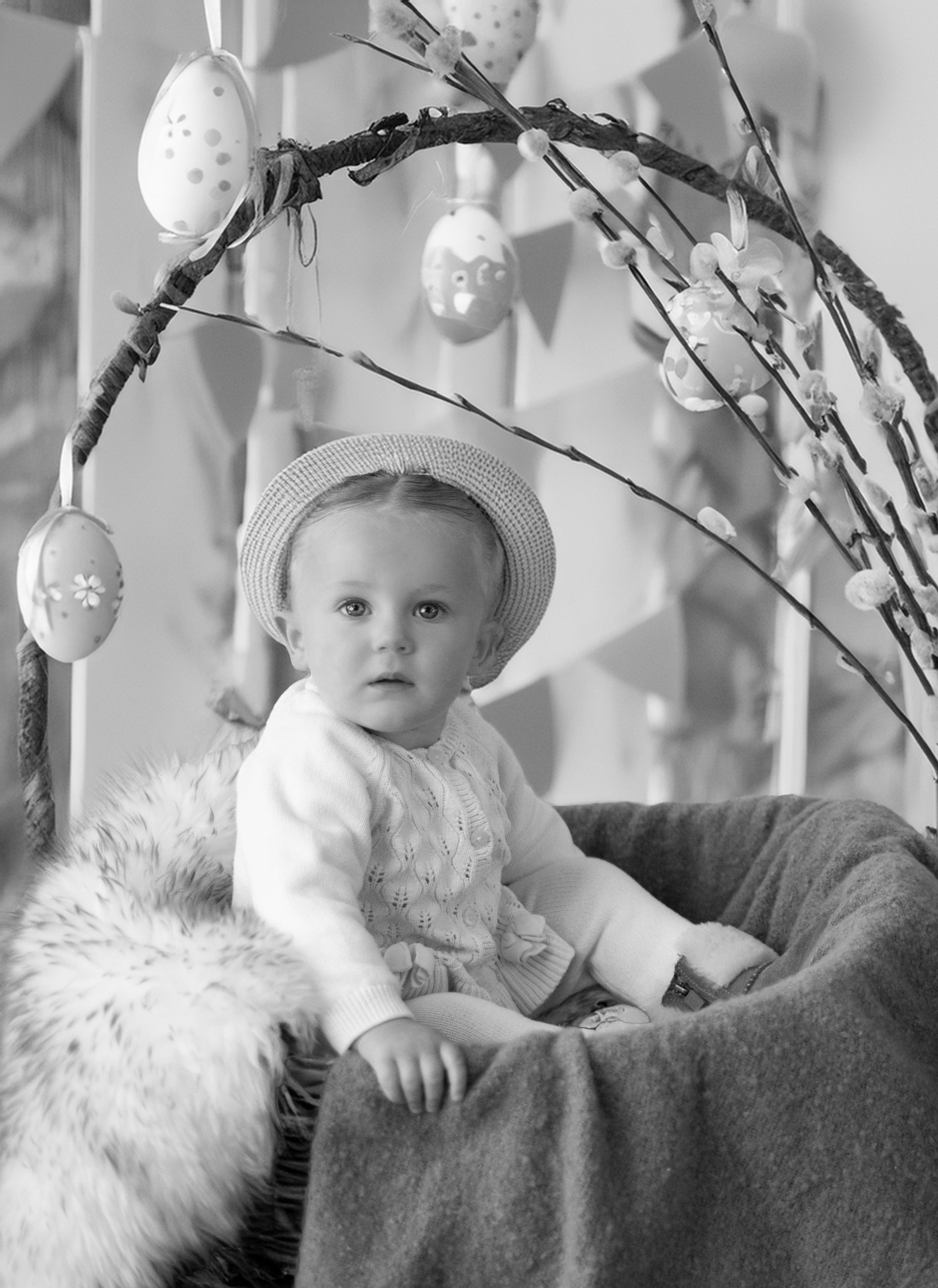 Vintage black and white Easter photograph showing a small child in a white sweater and hat seated with hanging egg decorations.