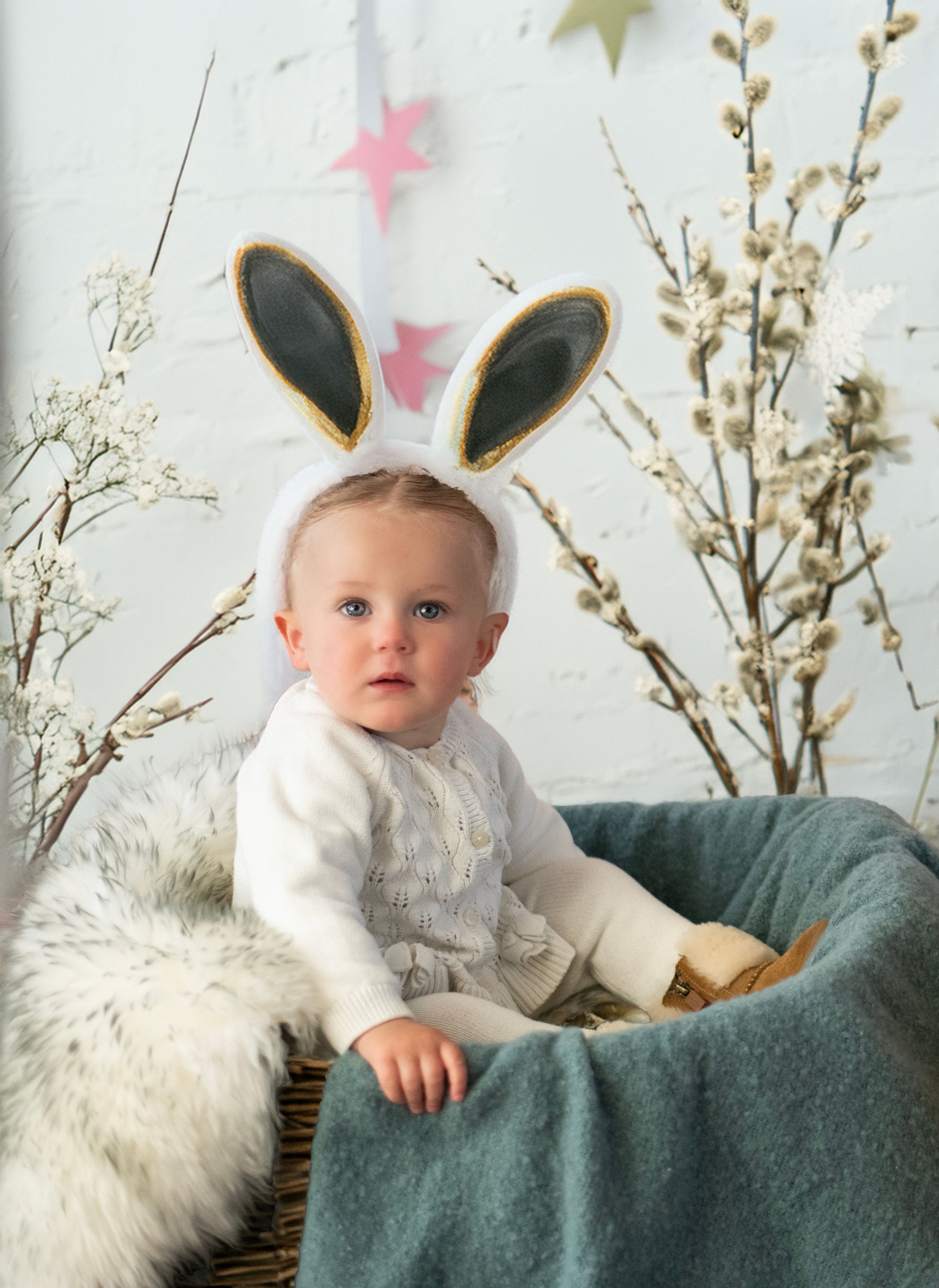 Small child in white sweater wearing bunny ears sits in grey chair with plush decor and spring branches in background.