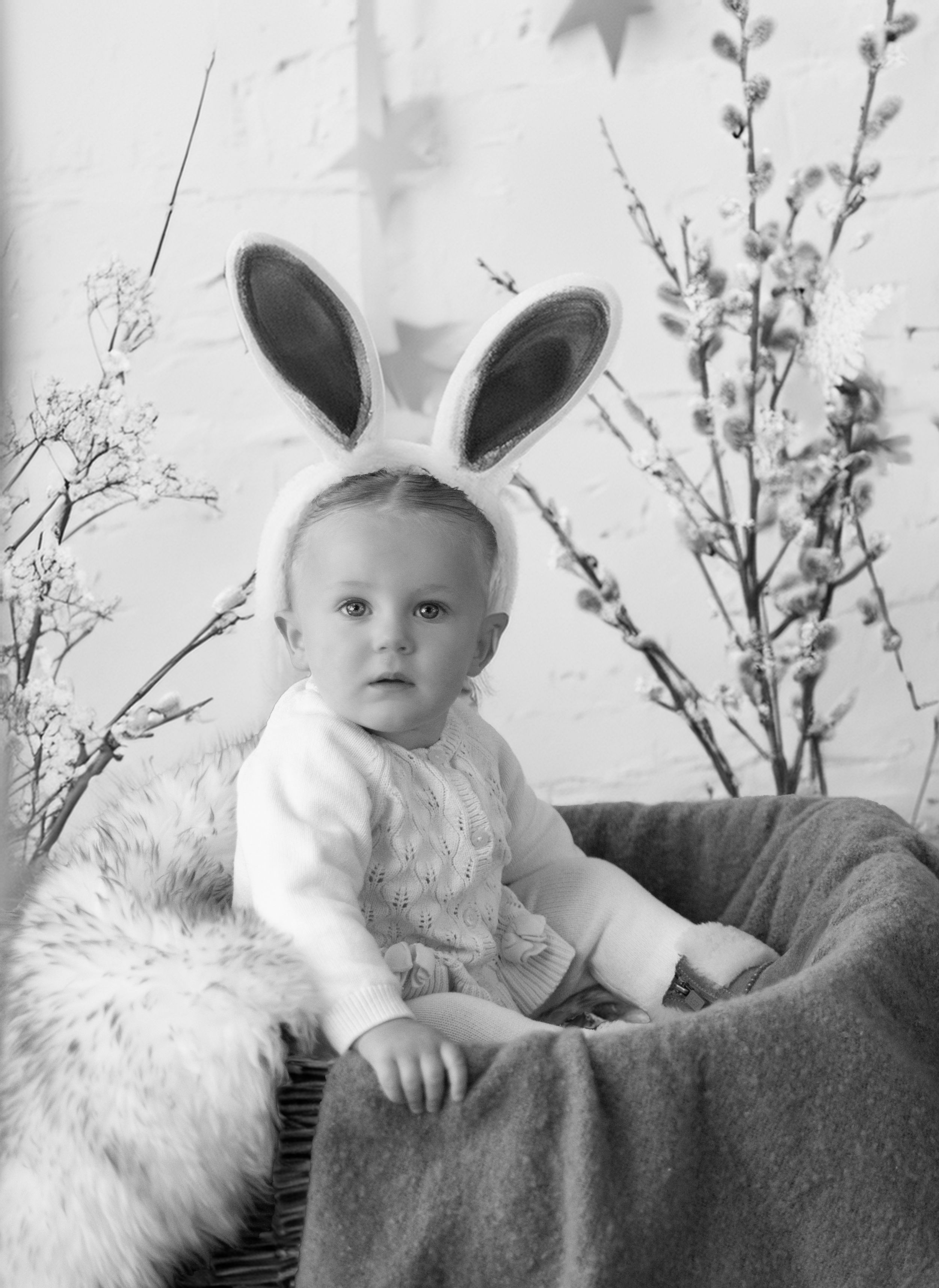 Black and white vintage photo shows small child wearing bunny ears while sitting on cushioned chair with spring decor.