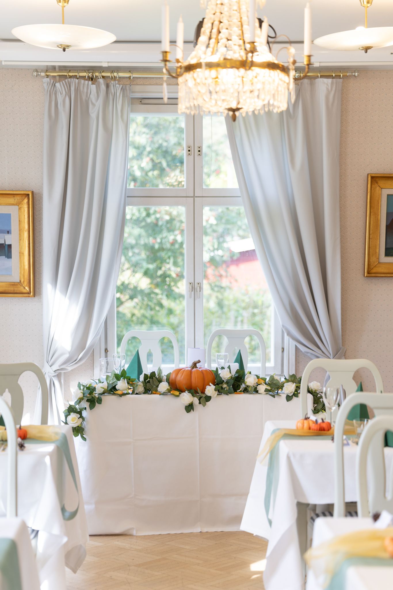 Elegant dining room with crystal chandelier, gray drapes, white tables, and autumn floral centerpiece with pumpkins near bright window.
