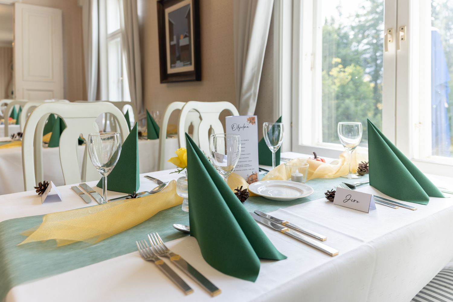 Elegant restaurant table setting with green folded napkins, white tablecloth, silverware, and wine glasses near bright windows.