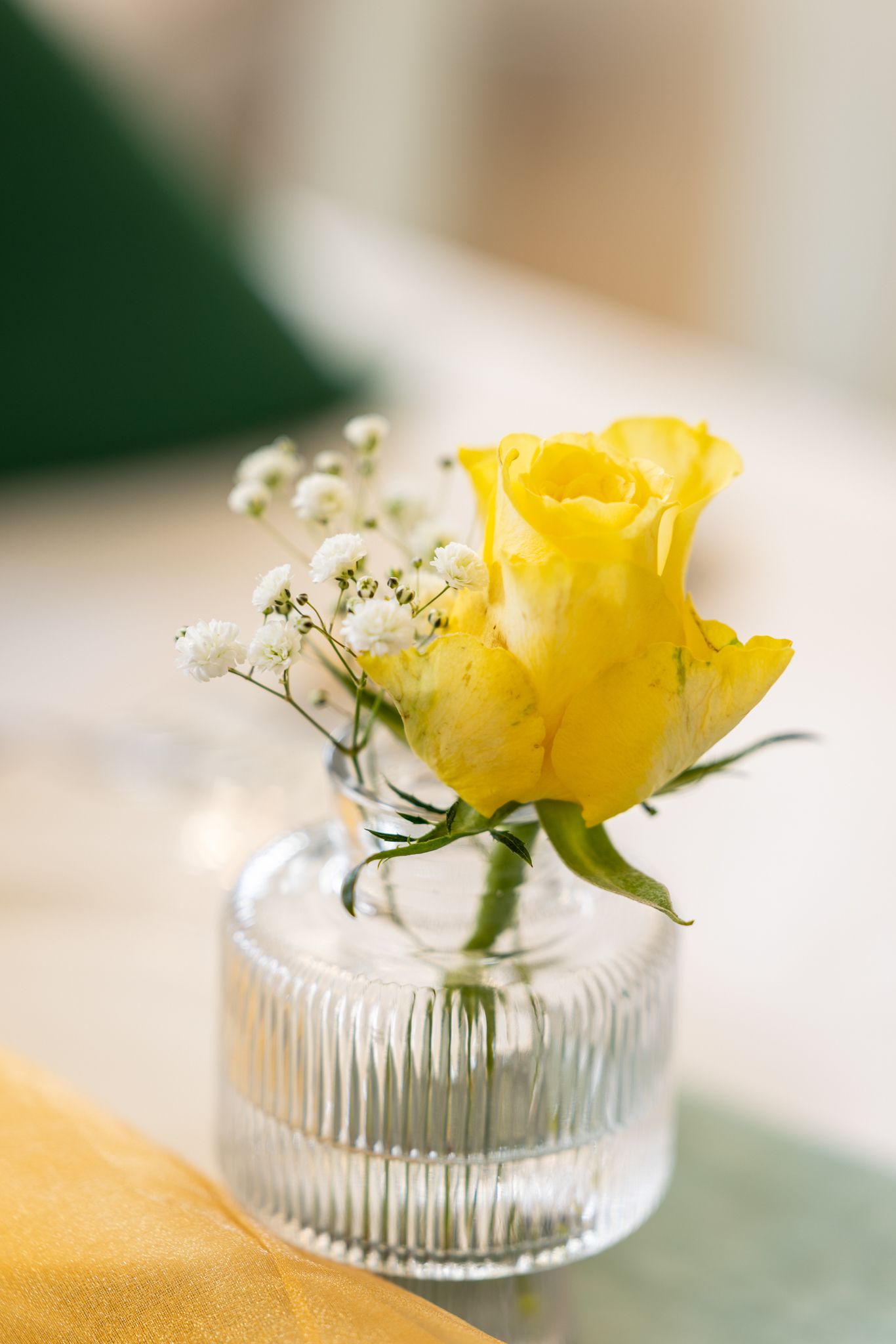 A vibrant yellow rose with baby's breath in a small ribbed glass vase on a table.