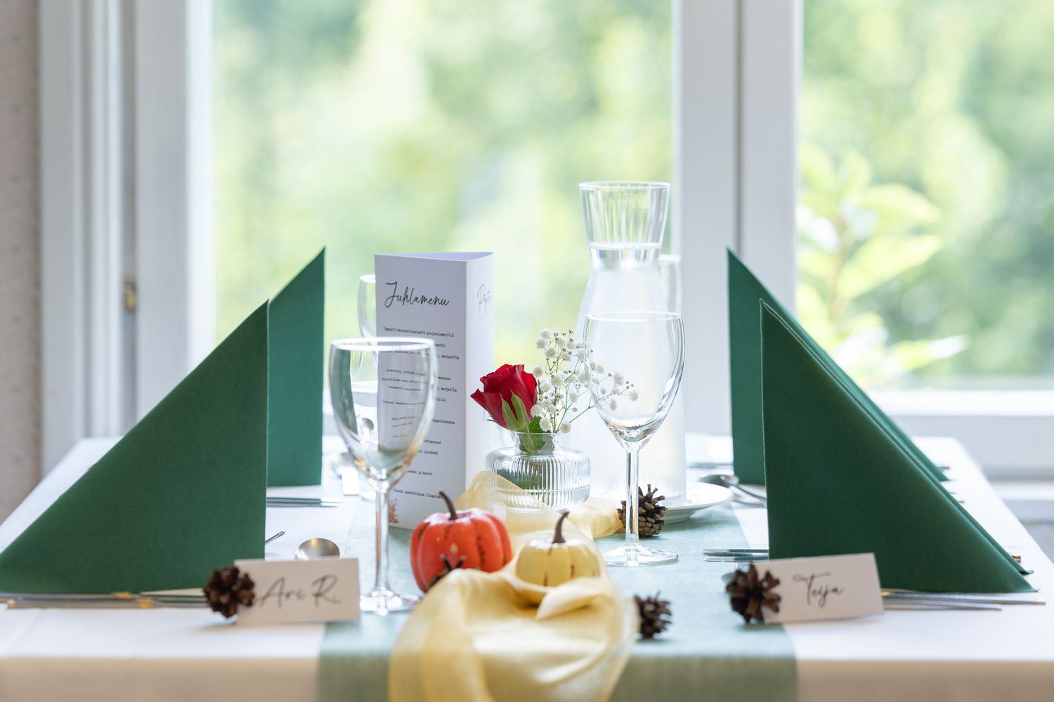 Elegant table setting with green folded napkins, wine glasses, mini pumpkins, pinecones, and place cards by a bright window.