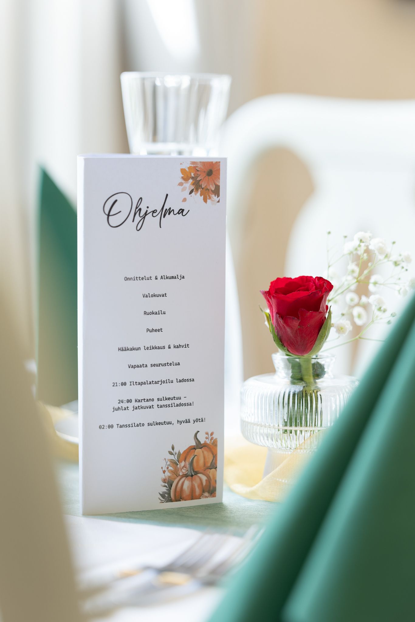 Elegant wedding program card with autumn pumpkin design beside a red rose in a glass vase on a decorated table.