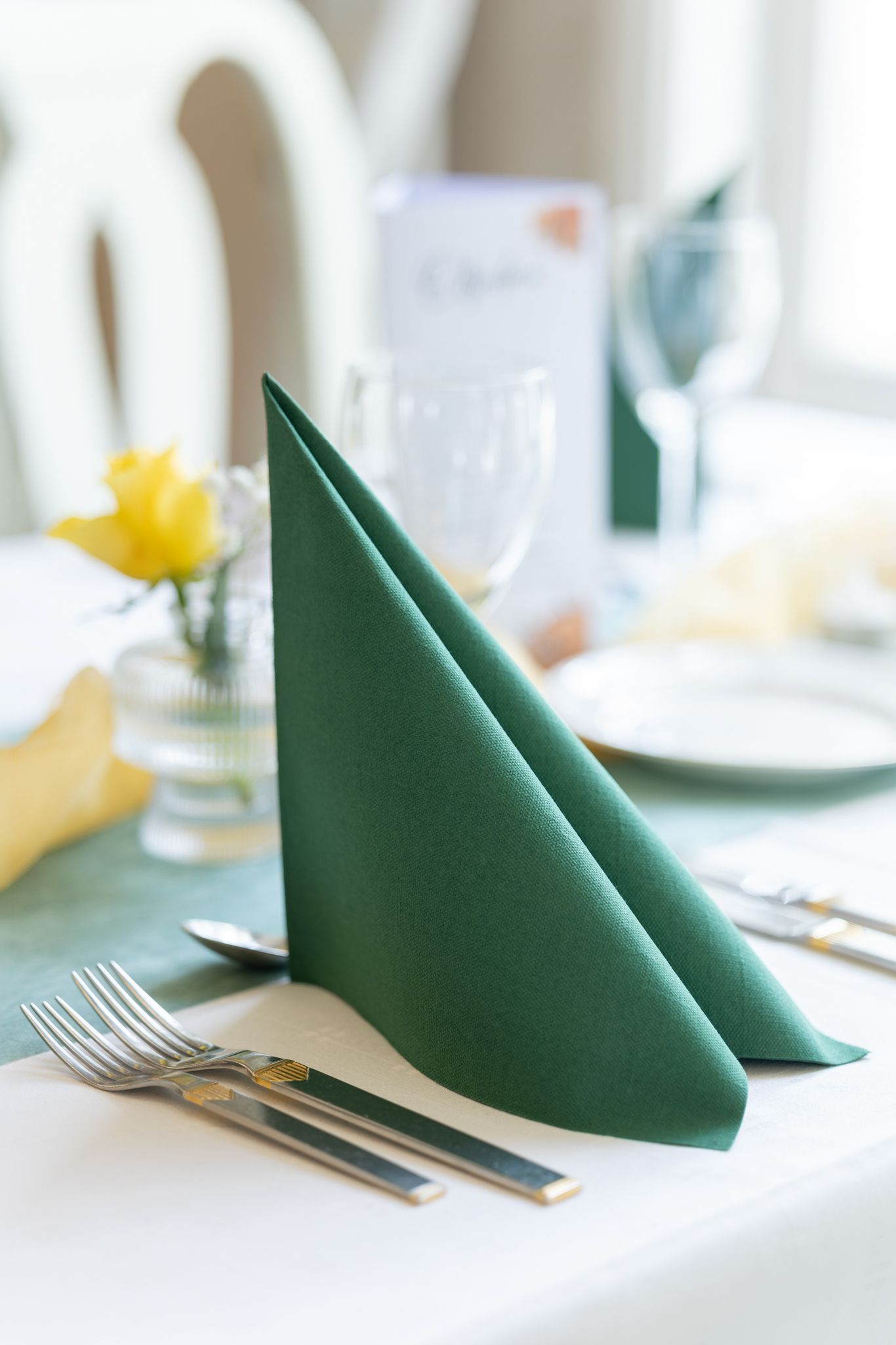Elegant green folded napkin on a formal dinner table setting with silverware, glassware, and yellow flowers.